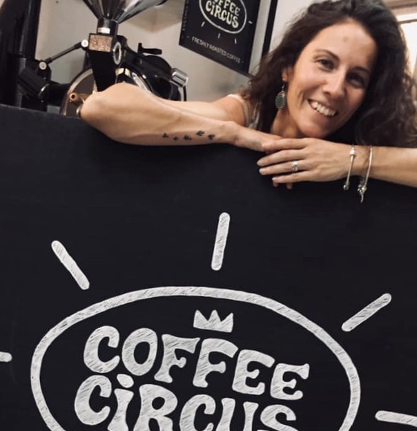 Coffee Circus Community member smiling behind hand-drawn Coffee Circus chalkboard sign with specialty coffee roasting equipment in background